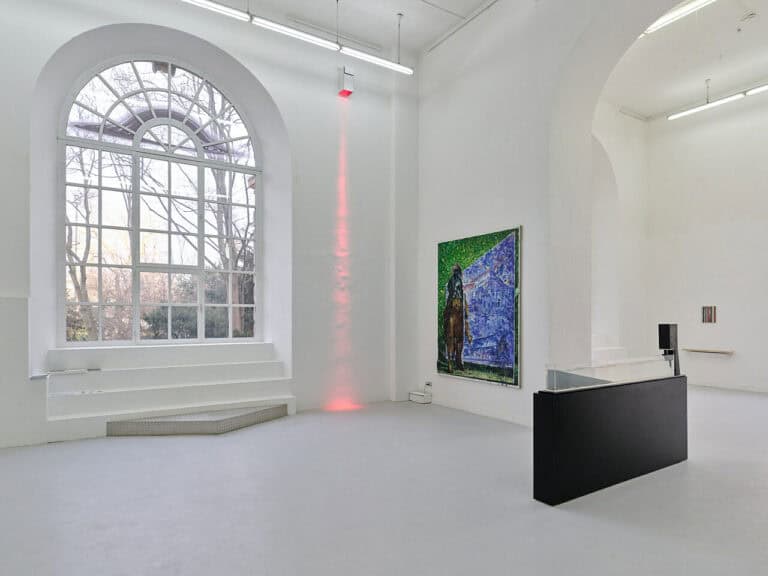 Foto: Ansicht eines Raumes mit weißen Wänden, grauem Boden und einem großen Bogenfenster. Im Raum und an den Wänden befinden sich mehrere Objekte in unterschiedlichen Formaten. Oben an einer der Wände ist ein Objekt angebracht, von dem ausgehend ein roter Lichtkegel sich über die Wand bis hin zum Boden erstreckt.