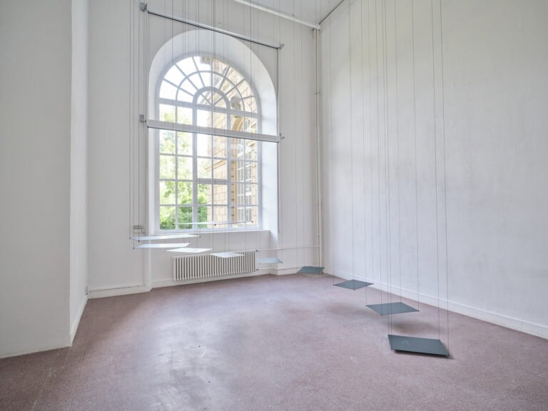 Foto: Ansicht eines Raumes mit weißen Wänden, rotem, gekacheltem Boden und einem großen Bogenfenster. Von der Decke hängen an Seilen mehrere Platten, die gestaffelt, in unterschiedlichen Höhen, über dem Boden schweben.