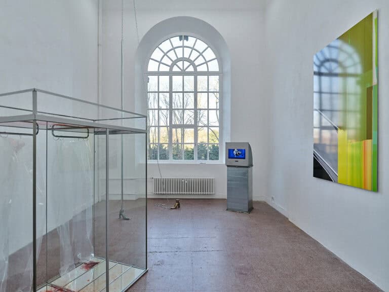 Foto: Ansicht eines Raumes mit rötlichem, steinernen Boden, weißen Wänden und einem großen Bogenfenster. Im Raum und an den Wänden befinden sich Objekte.
