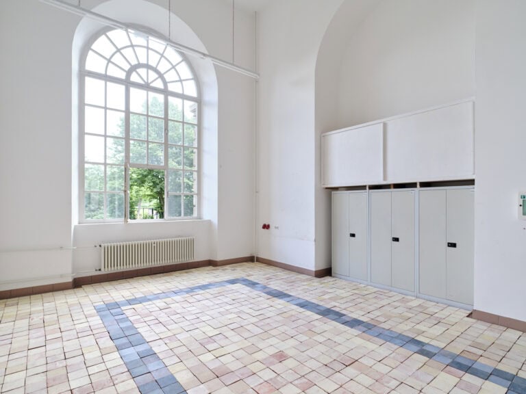 Foto: Ansicht eines Raumes mit weißen Wänden und einem großen Bogenfenster. In einer bogenförmigen Einlassung der Wände befinden sich Schränke. Der Boden des Raumes ist mit verschiedenfarbigen Kacheln ausgelegt.