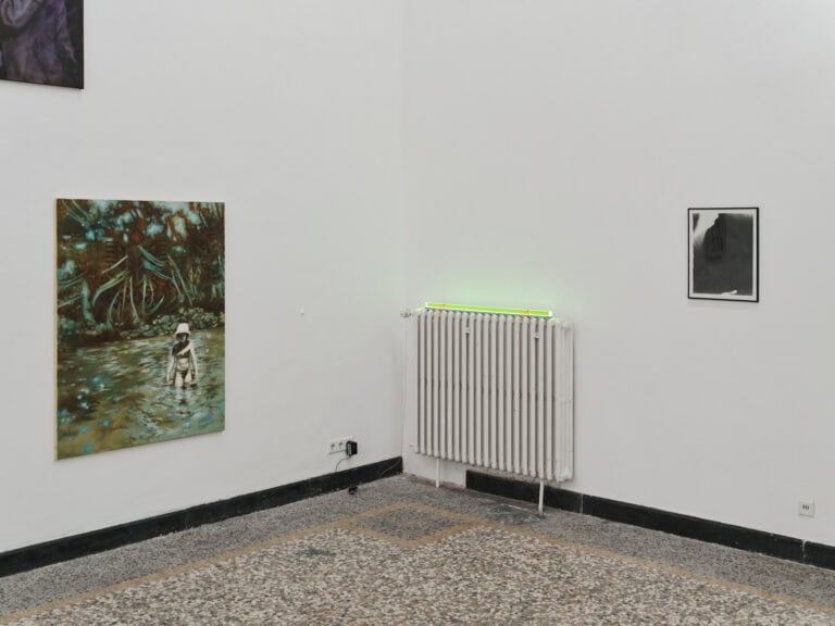 Foto: Ansicht eines Raumes mit weißen Wänden, einer schwarzen Fußleiste, einem mehrfarbigen Steinboden und einer weißen Heizung. Auf der Heizung liegt ein länglicher, leuchtender, neongrüner Gegenstand. An den Wänden befinden sich Objekte.