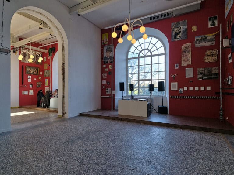 Foto: Ansicht eines Raumes mit mehrfarbigen Steinboden, roten und weißen Wänden und zwei Fenstern. In der Mitte des Raumes ist ein bogenförmiger Durchbruch der Wand zu sehen, der die Räume miteinander verbindet. Vor beiden Fenstern befindet sich eine niedrige Tribüne. Vor einem der Fenster ist eine Theke aufgebaut, um die herum Menschen stehen. Vor dem anderen Fenster steht ebenfalls eine Theke, die von vier Lautsprechern umgeben ist. Die Roten Wände befinden sich nahe der Fenster und sind bis unter die Decke mit Arbeiten behangen.