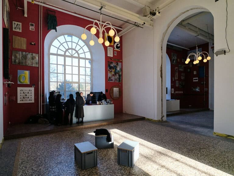 Foto: Ansicht eines Raumes mit mehrfarbigen Steinboden, roten und weißen Wänden und zwei Fenstern. In der Mitte des Raumes ist ein bogenförmiger Durchbruch der Wand zu sehen, der die Räume miteinander verbindet. Vor beiden Fenstern befindet sich eine niedrige Tribüne. Vor einem der Fenster ist eine Theke aufgebaut, um die herum Menschen stehen. Im Raum befinden sich außerdem drei Hocker. Die Roten Wände sind bis unter die Decke mit Arbeiten behangen.