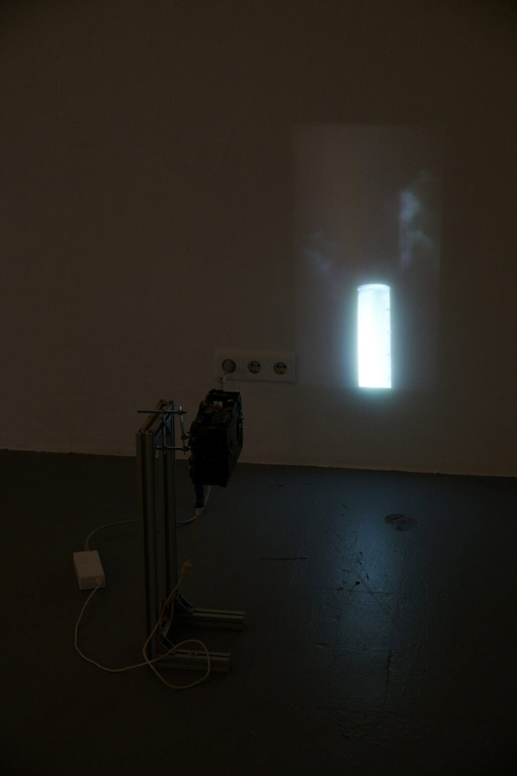 Foto: Ein abgedunkelter Raum. Im Raum steht wandnah ein kleines Gerüst, an dem ein Beamer angebracht ist. Der Beamer wirft eine Projektion an die Wand.