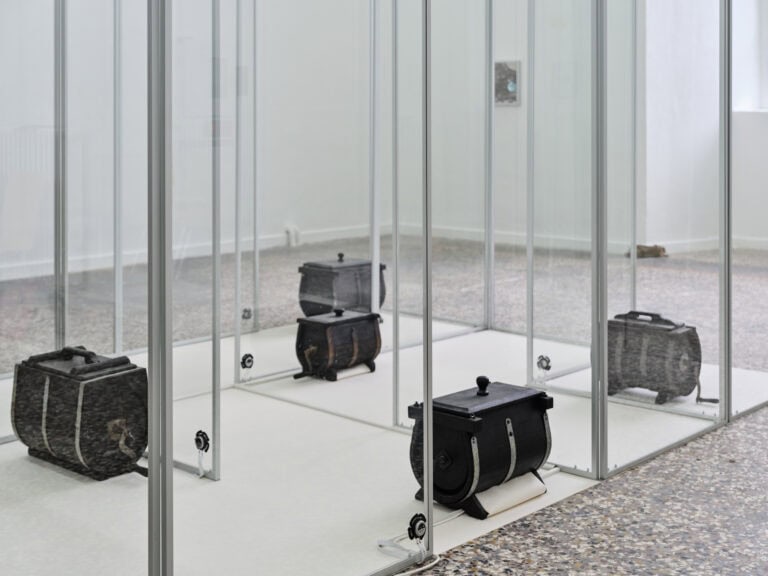 Foto: Nahaufnahme von schwarzen Boxen in einer Konstruktion aus Glasscheiben. Die Boxen stehen auf einem hellen Material. Die Wände des die Konstruktion umgebenden Raumes sind weiß, der Boden besteht aus mehrfarbigem Stein.