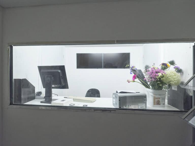 Foto: Nahaufnahme eines weißen Wand mit eingelassener, leicht milchiger Scheibe. Durch die Scheibe ist ein Raum zu erkennen, in dem sich Objekte, darunter mehrere Bildschirme, eine Tastatur, ein Drucker, Blumen und Hefte befinden.