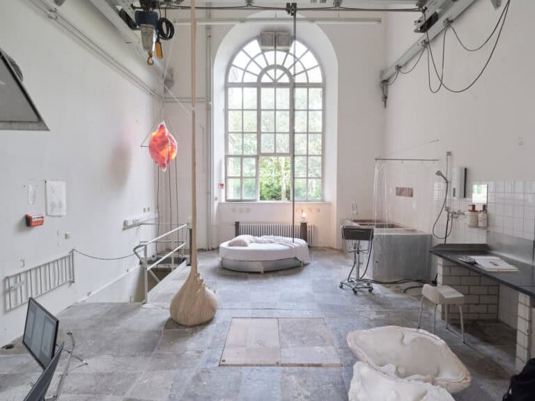 Foto: Ansicht eines Raumes mit weißen Wänden, grauem Boden und einem großen Bogenfenster. Im Raum befinden sich mehrere Objekte und Einrichtungsgegenstände einer Werkstatt.