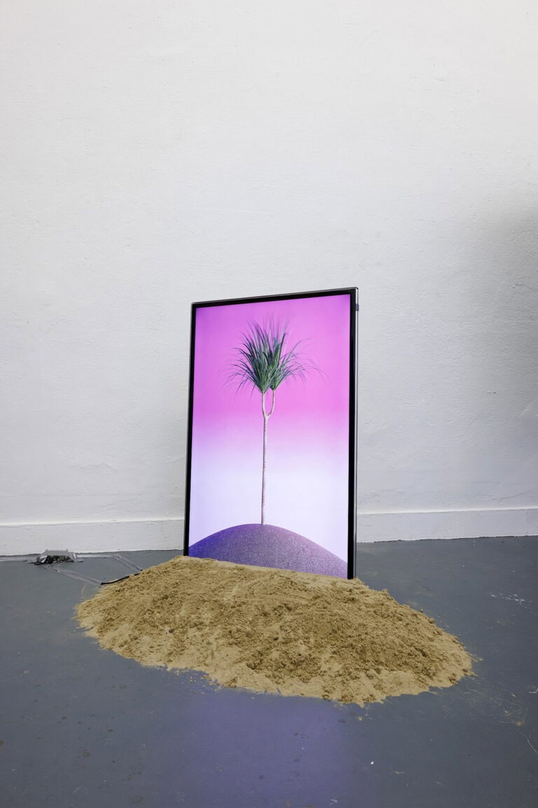 Foto: Aufnahme eines Objektes auf grauem Boden vor einer weißen Wand. Ein Bildschirm. der in einen Haufen Sand gesteckt ist.
