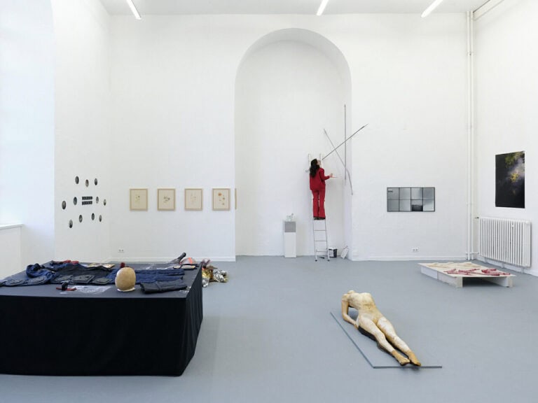 Foto: Ansicht eines Raumes mit weißen Wänden, grauem Boden und einer deckenhohen, bogenförmigen Einbuchtung. Im Raum befindet sich eine Vielzahl von Objekten, die in Teilen an der Wand montiert sind, auf Sockeln stehen oder auf dem Boden liegen. In der Einbuchtung steht eine Person auf einer Leiter.