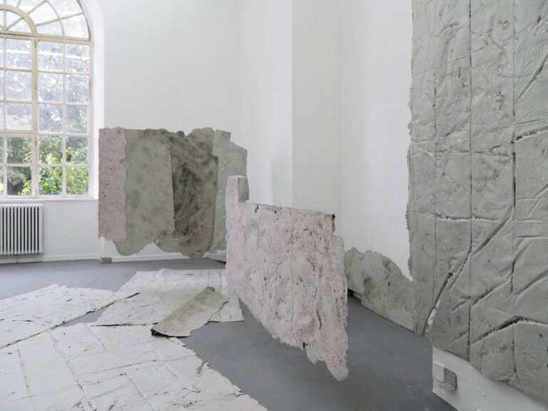 Foto: Ansicht eines Raumes mit weißen Wänden, grauem Boden und einem großen Bogenfenster. An den Wänden, auf dem Boden und frei im Raum von der Decke schwebend befinden sich mehrere flache, gräuliche Objekte.