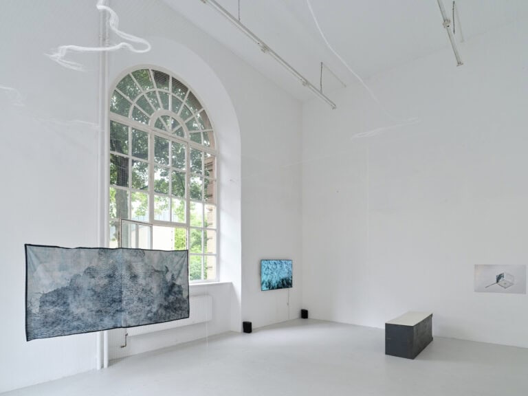 Foto: Ansicht eines Raumes mit weißen Wänden, grauem Boden und einem großen Bogenfenster. An den Wänden des Raumes, von der Decke hängend und im Raum befinden sich mehrere Objekte aus unterschiedlichen Materialien.