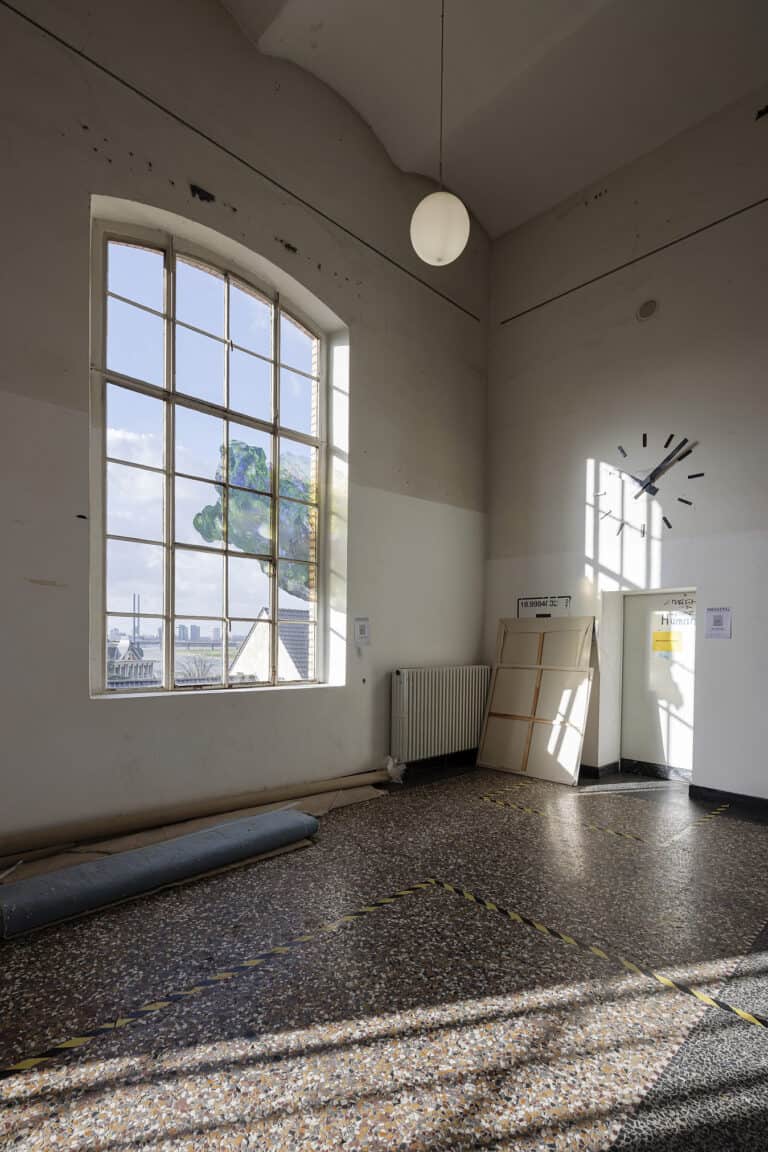 Foto: Ansicht einer Raumecke mit großem Bogenfenster. Sonnenlicht fällt durch das Fenster. Im Flur stehen Objekte und Leinwände, deren Rückseite zu sehen ist.
