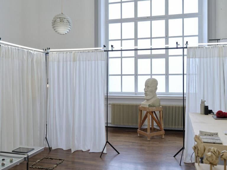 Foto: Ansicht eines Raumes mit weißen Wänden, großem Fenster und braunem Dielenboden. Im Raum befindet sich eine Konstruktion, an der weiße vorhänge befestigt sind, die auf den Boden reichen. Im Raum, vor und hinter den Vorhängen, befinden sich Objekte die auf Sockeln und Tischen angebracht sind oder auf dem Boden liegen.