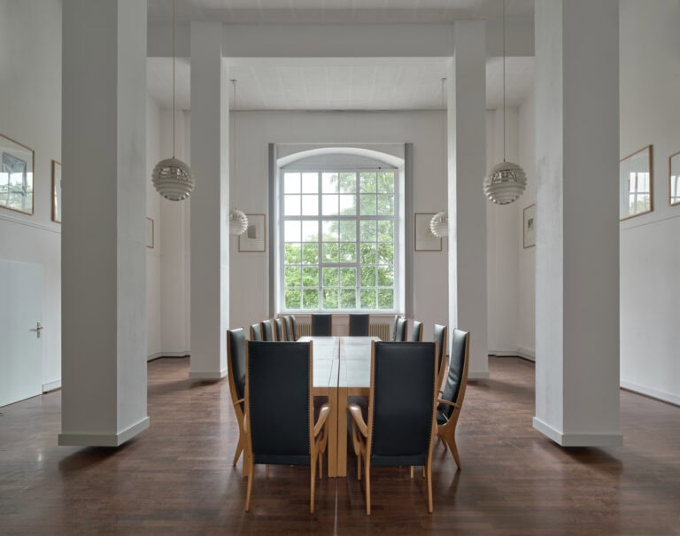 Foto: Ansicht eines Raumes mit weißen Wänden, vier weißen Säulen, die über dem Boden schweben und einem großen Bogenfenster. Von der Decke des Raumes reichen vier Lampen in den Raum hinein. An den Wänden hängen an der oberen hälfte der Wand mehrere gerahmte Objekte. In der Mitte des Raumes steht eine lange Tafel mit mehreren Stühlen mit hohen Lehnen.