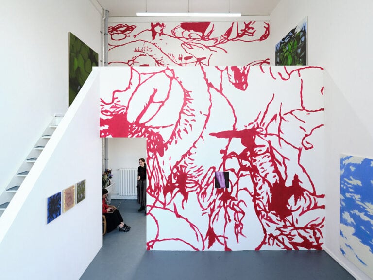 Foto: Ansicht eines Raumes mit weißen Wänden und grauem Boden. Im Raum befindet sich eine durch eine Treppe begehbarer Zweite ebene. Auf Teile der Wände ist mit roter Farbe ein Motiv aufgebracht. Unten und oben im Raum, teilweise auf den weißen Wänden, teilweise auf den Wänden die mit der roten Farbe bearbeitet wurden, hängen Leinwände im Klein-, Mittel- und Großformat. Im Raum befinden sich zwei Personen.