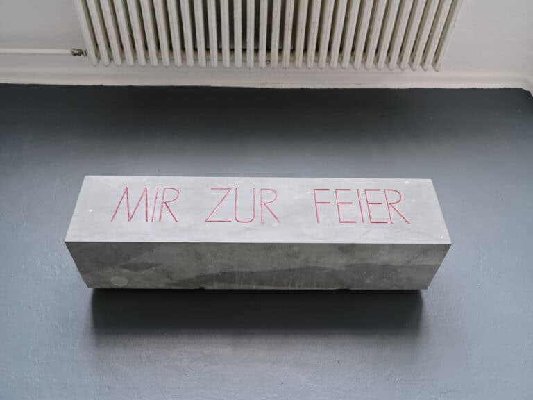 Foto: Aufnahme eines Objektes das auf grauem Boden liegt. Ein grauer Quader mit der Aufschrift "MIR ZUR FEIER".