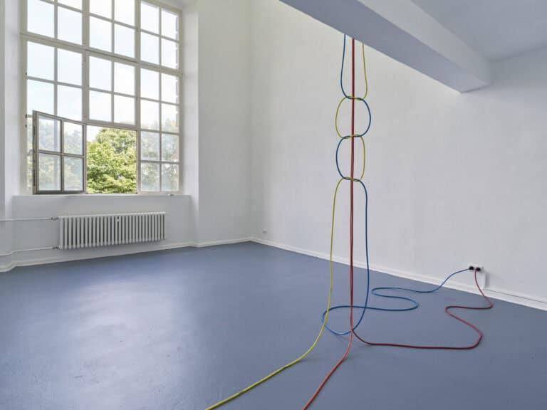 Foto: Ansicht eines Raumes mit weißen Wänden, blauem Boden und einem großen Fenster. Von der Decke hängen drei Farbige Seile, die miteinander verwoben sind. Zwei der Seile sind in eine Steckdose eingesteckt.