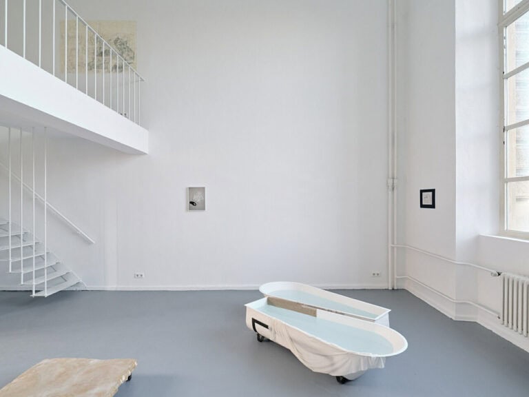 Foto: Ansicht eines Raumes mit weißen Wänden, grauem Boden und einem Fenster. An den Wänden und auf dem Boden des Raumes befinden sich unterschiedliche Objekte. Links im Bild ist ein Treppenaufgang zu sehen.