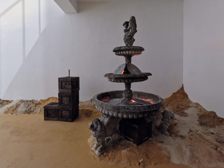 Foto:Ansicht eines Raumes mit weißen Wänden. Der Boden des Raumes ist mit gelblichem Sand bedeckt, der unterschiedlich hoch aufgeschüttet ist. Im Raum befinden sich ein steinerner Brunnen, in dem schwarze, runde Platten liegen hinter denen ein rötliches Licht hervortritt. Neben den Brunnen steht ein weiteres Objekt.
