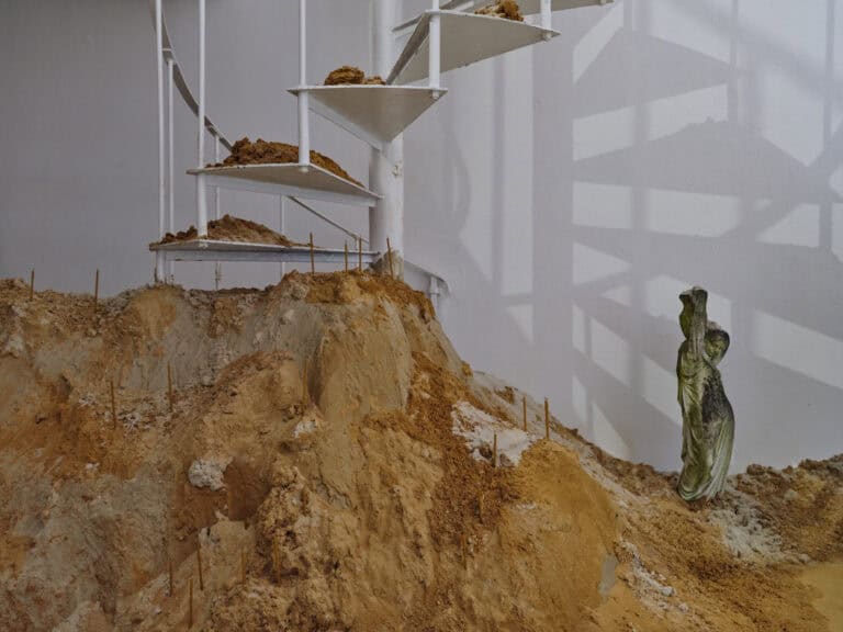 Foto: Ansicht eines Raumes mit weißen Wänden und einer Wendeltreppe. Auf den Treppenstufen und im Raum befindet sich eine große Menge von gelbem Sand. In den Sand sind schmale gelbe Kerzen und eine moosbewachsene Skulptur gesteckt.