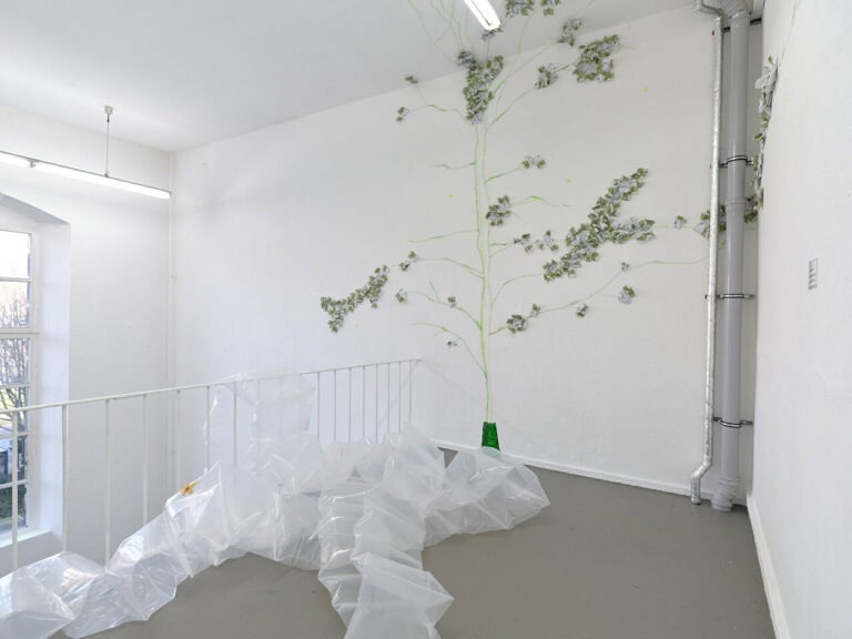 Foto: Ansicht eines Raumes mit weißen Wänden, grauem Boden und einem großen Bogenfenster. In der Ecke des Raumes befinden sich graue Rohe. Mittig im Raum befindet sich ein weißes Geländern. Am Boden des Raumes und an den Wänden befinden sich Objekte.