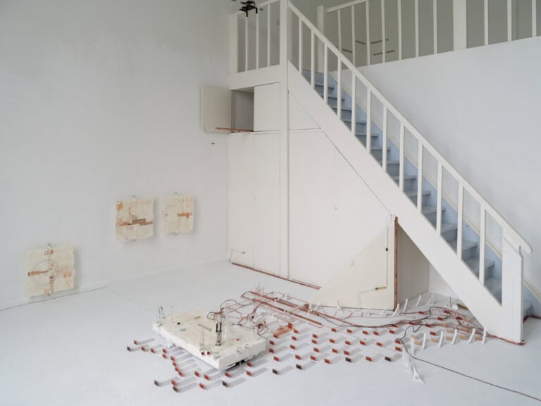 Foto: Ansicht eines Raumes mit weißen Wänden, weißem Boden und einer Treppe mit weißem Geländer, die zu einer weiteren Ebene im Raum führt. Auf dem Boden des Raumes, an den Wänden und an dem Geländer installiert befinden sich mehrere Objekte.