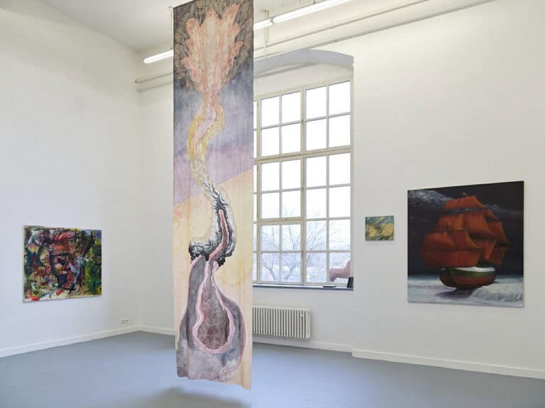 Foto: Ansicht eines Raumes mit weißen Wänden, grauem Boden und einem großen Bogenfenster. Im Raum hängen Malereien auf unterschiedlichen Höhen und in unterschiedlichen Formaten. Von der Decke hängt ein mit Farbe bearbeitetes Textil bis kurz über den Boden.