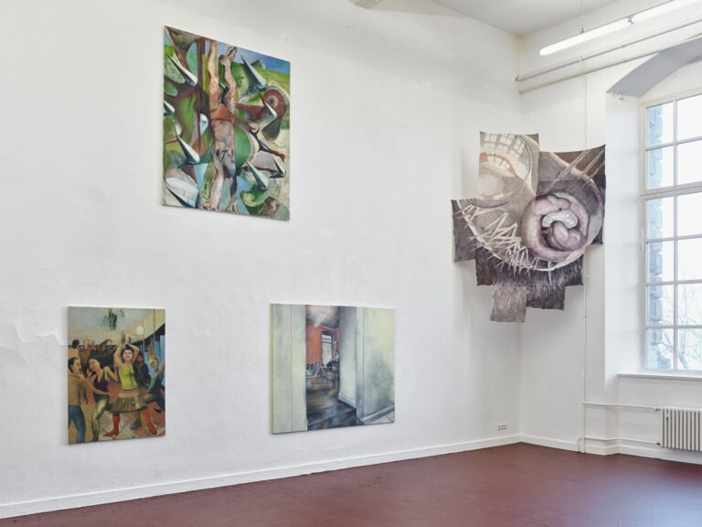 Foto: Ansicht eines Raumes mit einem großen Fenster, weinrotem Boden und einer weißen Wand. An der Wand hängen auf unterschiedlichen Höhen Bilder in großen Formaten.