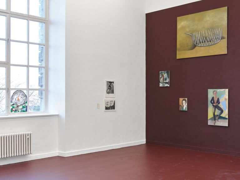 Foto: Ansicht eines Raumes mit einem Fenster, einer weißen Wand, einer lila Wand und einem weinroten Boden. Vor dem Fenster und an den Wänden befinden sich Objekte und Malereien in Klein-, Mittel- und Großformaten.