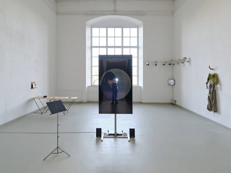 Foto: Ansicht eines Raumes mit weißen Wänden, grauem Boden und einem großen Bogenfenster. An den Wänden und im Raum stehen unterschiedliche Objekte.