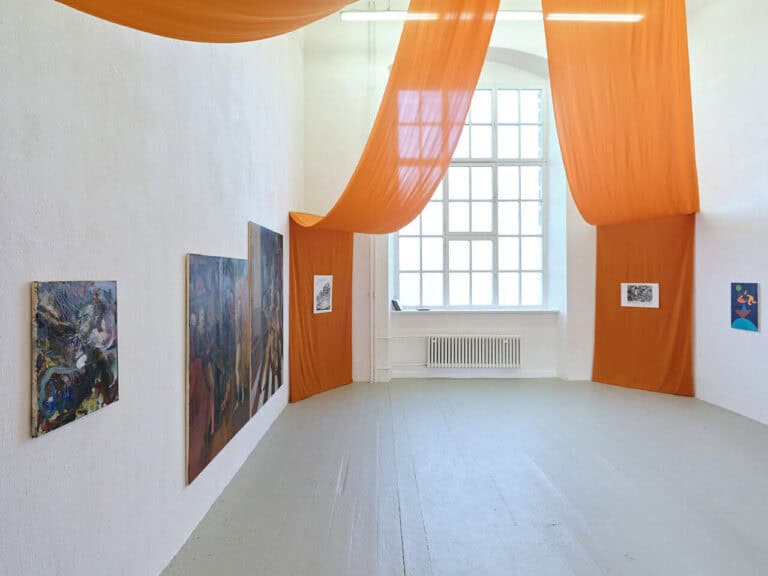 Foto: Ansicht eines Raumes mit weißen Wänden, grauem Dielenboden und einem Fenster. Von der Decke reichen drei orangene Tücher an die Wände bis zum Boden. Auf den Wänden hängen Arbeiten im Klein-, Mittel- und Großformat.