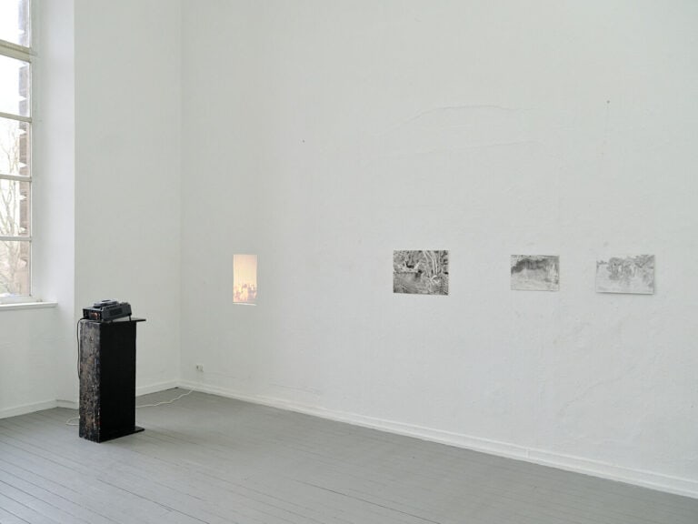 Foto: Ansicht eines Raumes mit weißen Wänden, grauem Dielenboden und einem Fenster. An der Wand befindet sich drei Papierarbeiten. Vor der Wand steht ein Beamer auf einem Sockel, der eine Projektion an die Wand wirft.