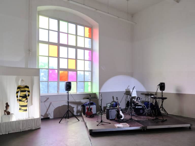 Foto: Ansicht eines Raumes mit weiß-grauen Wänden, grauem Boden und einem Fenster. Die Fensterscheiben sind teilweise mit bunten Folien beklebt. im Raum befindet sich ein mit Stoff ausgekleideter Schaukasten, in dem sich eine bekleidete Schaufensterpuppe und Schuhe befinden, die auf einem Podest stehen. Daneben ist eine Bühne installiert, auf der sich Instrumente und Technik befinden.
