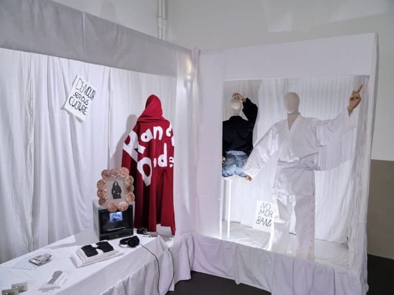 Foto: Ansicht einer Raumecke mit weißen Wänden und dunklem Boden. Im Raum ist ein Schaukasten installiert, der mit weißem Stoff ausgehangen ist. Im Schaukasten befinden sich bekleidete Schaufensterpuppen, Objekte und beschriebene Papiere.