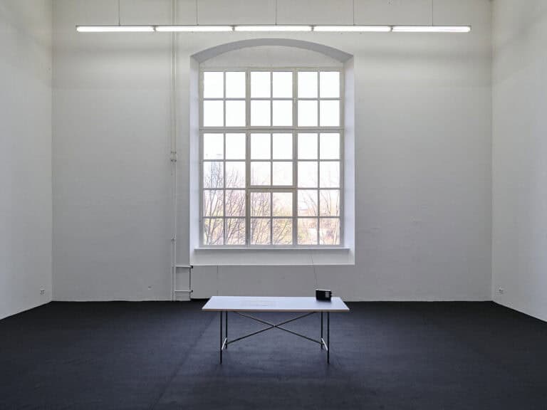 Foto: Ansicht eines Raumes mit weißen Wänden, dunklem Boden und einem großen Bogenfenster. In der Mitte des Raumes ist ein Tisch platziert, auf dem ein Radio steht.