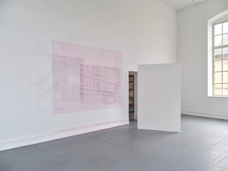 Foto: Ansicht eines Raumes mit weißen Wänden, grauem Dielenboden und einem Fenster. Die Wand wurde direkt mit grauer und Rosner Farbe bearbeitet. Im Raum steht eine schmale, weiße Trennwand.