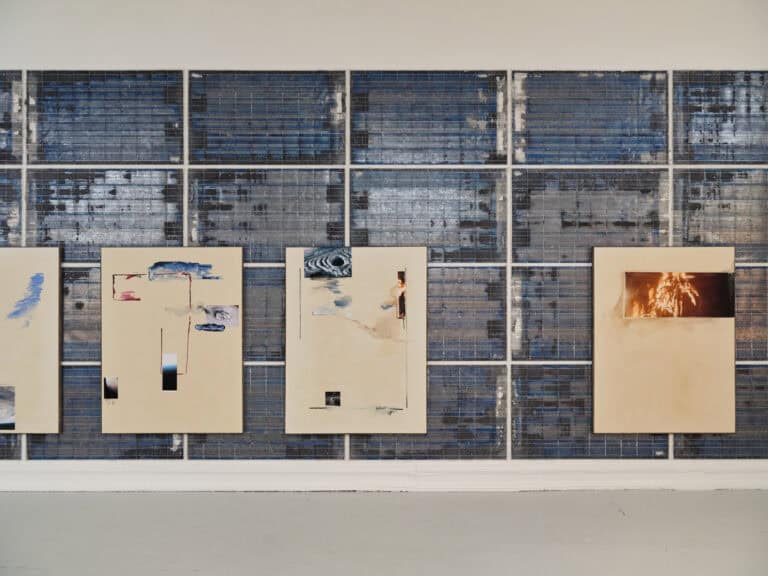 Foto: Ansicht eines Raumes mit weißer Wand und grauem Boden. Ein Großteil der Wand ist von glänzenden Banalen Bedeckt, auf denen Malereien angebracht sind.