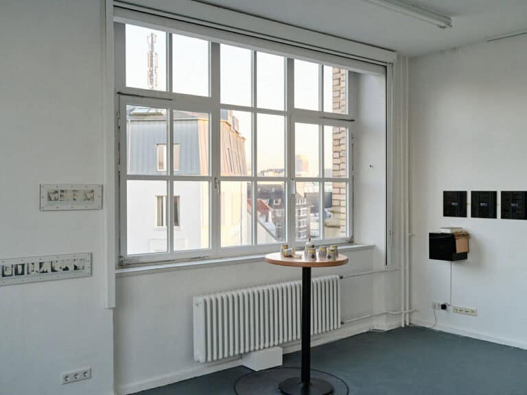 Foto: Ansicht einer Raumecke mit weißen Wänden, dunklem Boden und einem Fenster. An den Wänden hängen Objekte. Vor dem Fenster steht ein kleiner, hoher, runder Tisch mit Papierbechern.