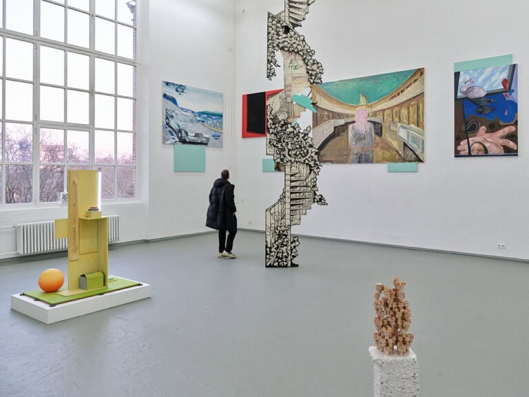 Foto: Ansicht eines Raumes mit weißen Wänden, grauem Boden und einem großen Fenster. Im Raum und an den Wänden befinden sich Objekte. Die Objekte an den Wänden sind hoch gehangen. Im Raum befindet sich eine Person.