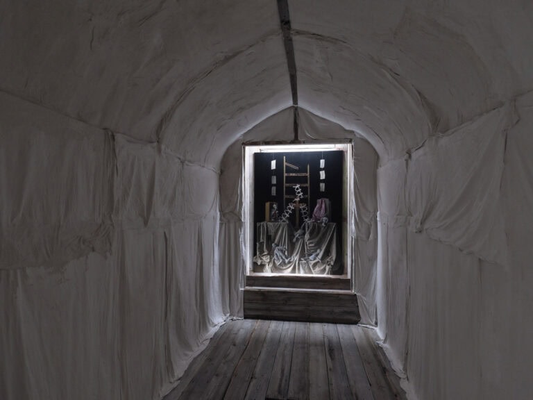 Foto: Ansicht eines schmalen Raumes, dessen Wände und Decke mit hellem Stoff abgehangen sind. Der Boden besteht aus hölzernen Dielen. Am Ende des Raumes ist eine Aussparung, in der sich eine großformatige Malerei befindet. Die Malerei wird von oben und unten von weißem Licht angestrahlt.