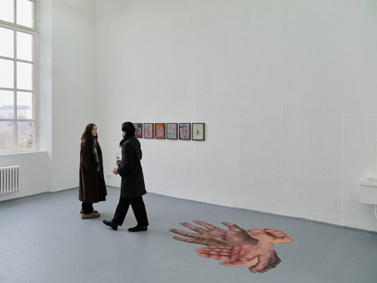 Foto: Ansicht eines Raumes mit weißen Wänden und grauem Boden. Auf dem Boden und an der Wand befinden sich Objekte. Im Raum bewegen sich zwei Personen.