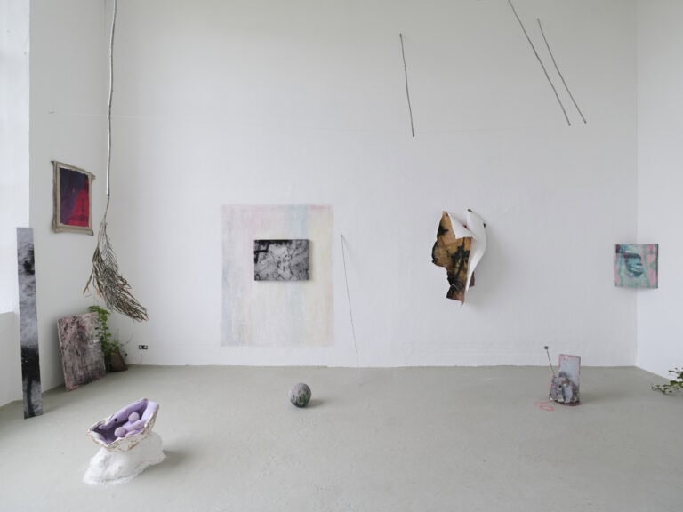 Foto: Ansicht eines Raumes mit weißen Wänden und grauem Boden. Auf dem Boden stehen eine Vielzahl von Objekten, manche von ihnen lehnen an der Wand. An de Wänden hängen, auf unterschiedlicher Höhe, weitere Objekte.