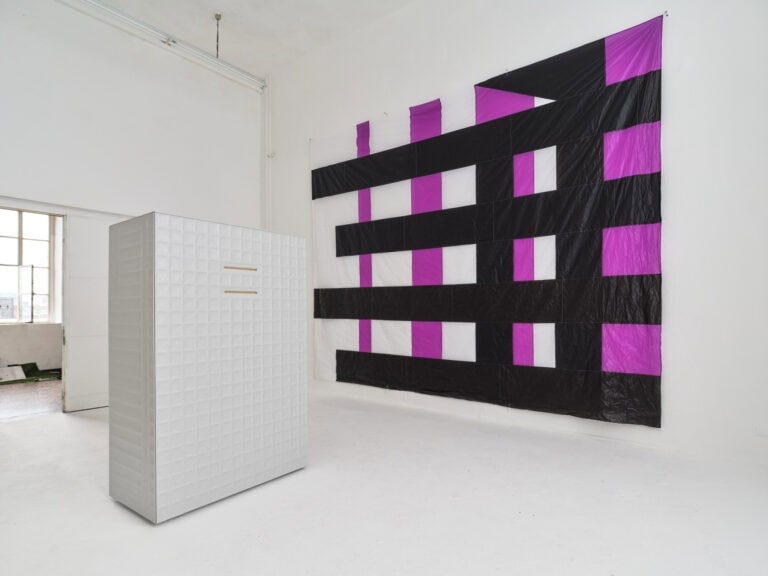 Foto: Ansicht eines Raumes mit weißen Wänden, weißem Boden und einer zweiflügligen, halb geöffneten Tür. An der Wand befindet sich ein großes, Flaches Objekt. Im Raum steht ein weiteres großes, quadratisches Objekt.