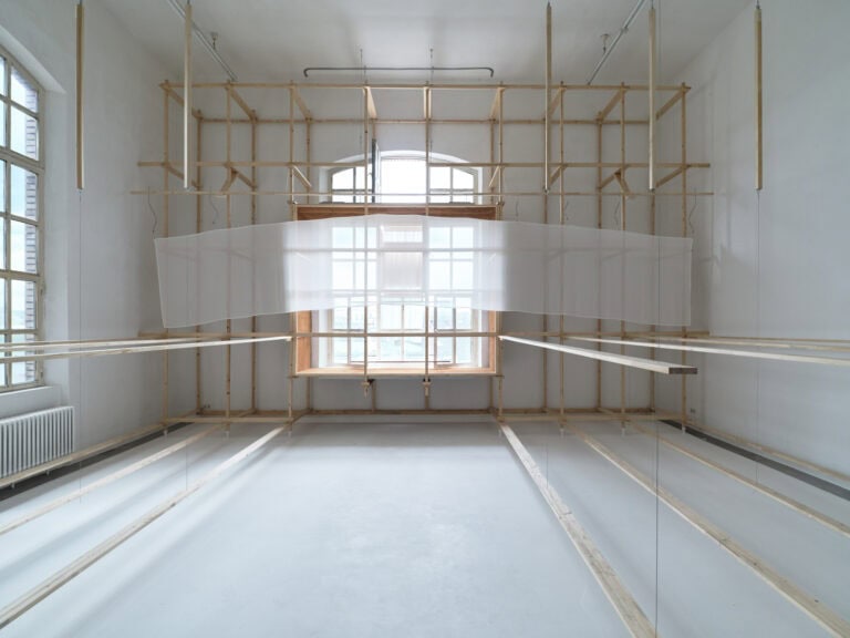 Foto: Aufnahme eines Raumes mit weißen Wänden, grauem Boden und zwei großen Bogenfenstern. Der Raum ist mit einer Konstruktion aus Holzlatten versehen, die sich durch die gesamte Höhe des Raumes erstreckt. Die Latten sind zum Teil durch Drähte miteinander verbunden und berühren den Boden nicht. Zwischen den Latten und Drähten ist ein halbtransparentes Textil gespannt.