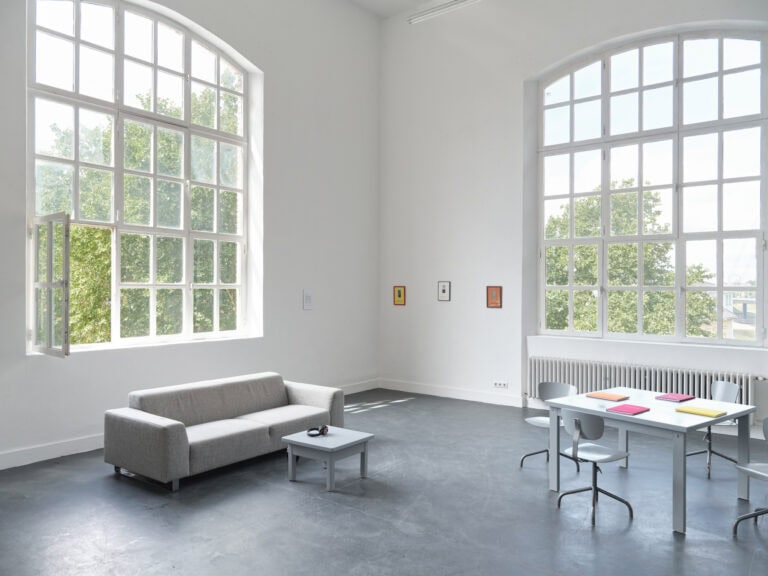 Foto: Ansicht eines Raumes mit weißen Wänden, grauem Boden und zwei großen Bogenfenstern. An den Wänden des Raumes hängen mehrere kleine Objekte. Im Raum verteilt befinden sich ein graues Sofa, ein kleiner grauer Tisch, ein großer grauer Tisch und vier graue Stühle. Auf den Tischen liegen Bücher und ein Kopfhörer.