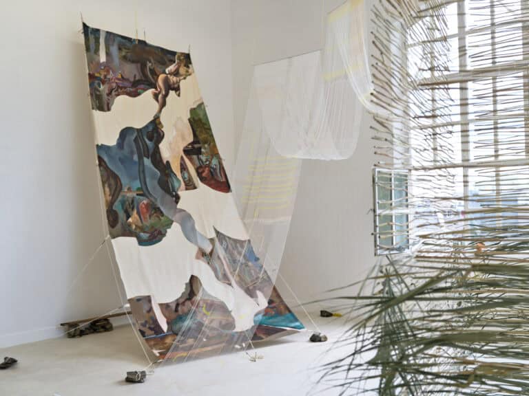 Foto: Ansicht eines Raumes mit weißen Wänden, beigem Boden und einem Fenster. Von der Decke hängen mehrere Textilien zum Boden herunter, eines von ihnen ist mit auf dem Boden liegenden Steinen beschwert.