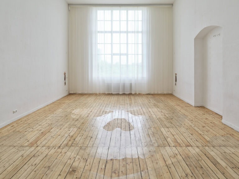 Foto: Ansicht eines Raumes mit weißen Wänden, hellem Dielenboden und einem großen Bogenfenster, vor dem lange, weiße, halbtransparente Vorhänge hängen, durch die das Licht fällt. Fensternah sind an der Wand gegenüber voreinander zwei Objekte auf gleicher Höhe angebracht. Teile des Bodens sind mit einem das Licht reflektierendem Material bedeckt.