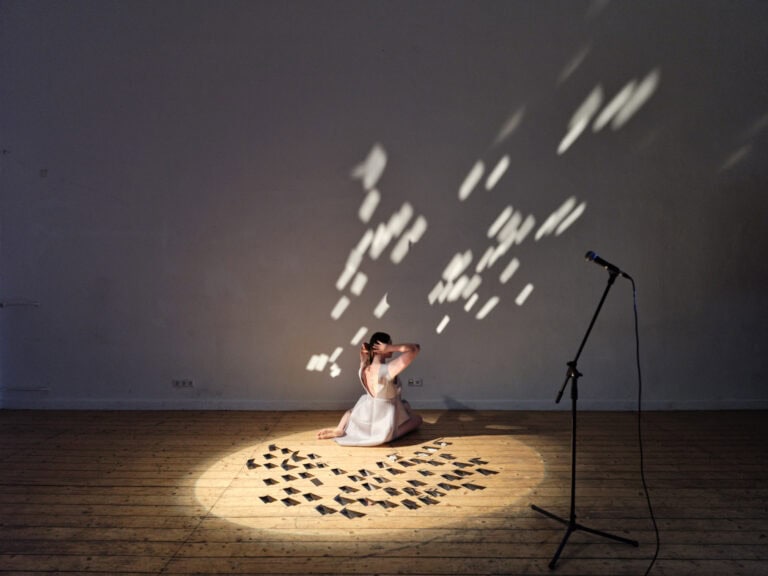 Foto: Ansicht eines abgedunkelten Raumes mit weißen Wänden und Dielenboden. Im Raum steht ein Mitkrofonständer. Auf dem Boden liegt eine Gruppe von Objekten, die von einem Lichtkegel angestrahlt wird. Das Licht wird von den Objekten reflektiert und wirft Schatten an die Wände. Zwischen der Wand und den Objekten sitzt ein Mensch mit angewinkelten Beinen auf dem Boden, der mit seinen Händen an den Hinterkopf fasst.