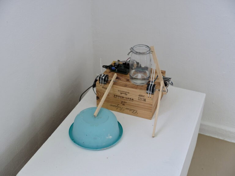 Foto: Nahausnahme von einer Vielzahl an Objekten aus unterschiedlichen Materialien auf einem weißen Sockel, vor einer weißen Wand.