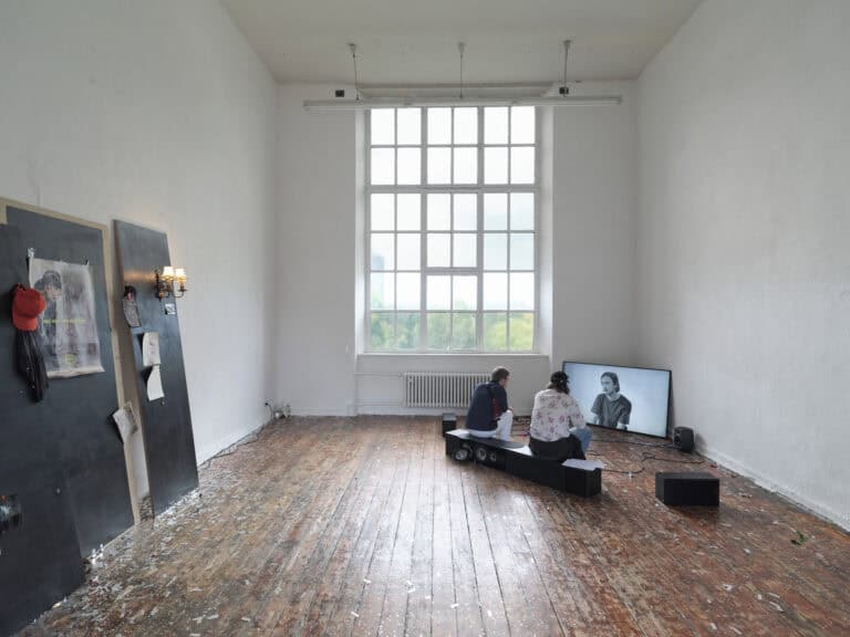 Foto: Ansicht eines Raumes mit weißen Wänden, großem Bogenfenster und braunem Dielenboden. An einer der Wände lehnen drei dunkle Platten an denen eine Lampe, Kleidung und Papiere hängen. In der rechten Ecke des Raumes stehen ein Bildschirm und mehrere Lautsprecher auf dem Boden. Auf einem der Lautsprecher sitzen zwei Menschen.